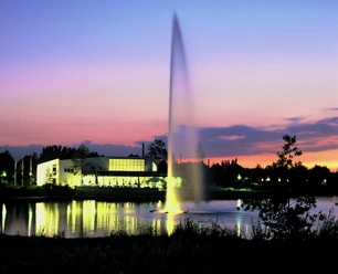 Seecafe_2.jpeg Ein großer Springbrunnen auf einem See im Vordergrund, beleuchtet bei Sonnenuntergang mit Gebäuden am Ufer.
