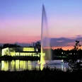 Seecafe_2.jpeg Ein großer Springbrunnen auf einem See im Vordergrund, beleuchtet bei Sonnenuntergang mit Gebäuden am Ufer.