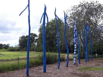 Blaue, schmale Skulpturen ragen dynamisch in den Himmel, umgeben von grünen Bäumen und Wiesen.