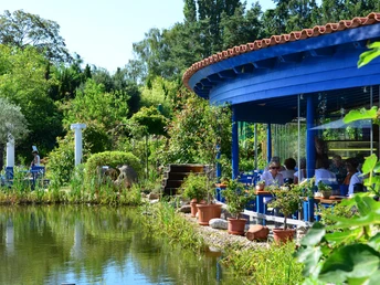 Rilaerleben Griechenland Eine blaue Terrasse mit Runddach überblickt einen Teich, umgeben von üppigem Grün und strahlender Sonne.