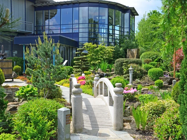 Japanischer Garten mit kleinen Brücke, umgeben von bunten Pflanzen und einem modernen Gebäude im Hintergrund.