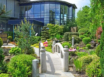 Rila erleben Japanischer Garten mit kleinen Brücke, umgeben von bunten Pflanzen und einem modernen Gebäude im Hintergrund.