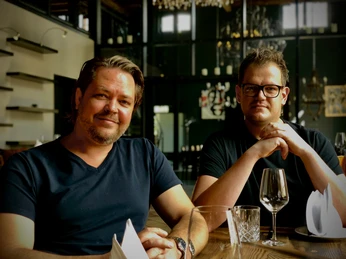 Abrahams_1.jpg Zwei Männer in einem Restaurant, die entspannt am Holztisch mit leeren Weingläsern sitzen.