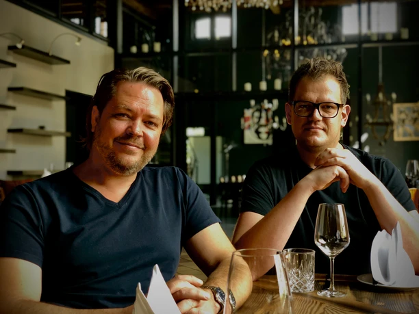 Abrahams_1.jpg Zwei Männer in einem Restaurant, die entspannt am Holztisch mit leeren Weingläsern sitzen.