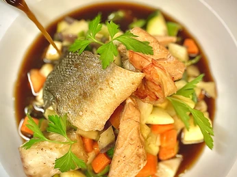 Gourmetgericht mit frischem Fisch, Garnelen und bunt gewürfeltem Gemüse in dunkler Sauce angerichtet.