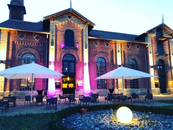 Historisches Backsteingebäude bei Dämmerung, farbig beleuchtet, mit Terrassenbestuhlung und Schirmen.