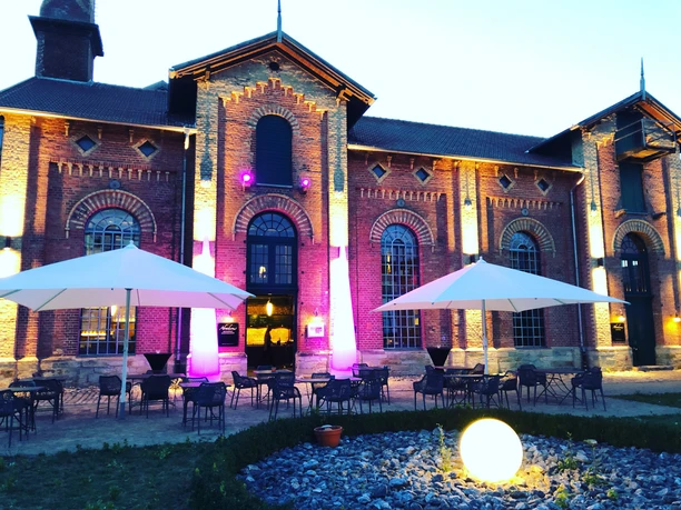 Historisches Backsteingebäude bei Dämmerung, farbig beleuchtet, mit Terrassenbestuhlung und Schirmen.