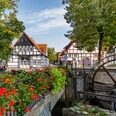 Mühlräder Nordwall Wiedenbrück Fachwerkhäuser mit roten Dächern umgeben eine historische Wassermühle, eingerahmt von bunten Blumen.