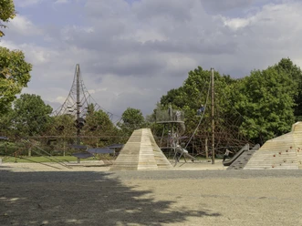 Moderner Spielplatz mit Kletterpyramiden, Balancierseilen und Spielgeräten, umgeben von Bäumen und Himmel.
