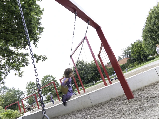 Schaukelparade an der Mittelhegge Ein Kind schaukelt glücklich auf einem Spielplatz mit roten Schaukelgestellen in einer grünen Umgebung.