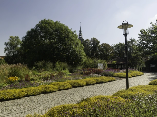 Ein gepflasterter Weg führt durch einen farbenfrohen Blumenpark mit Lampe und Kirchturm im Hintergrund.