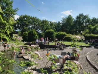 Eine sonnige Parklandschaft mit Personen beim Entspannen und Spielen in einem naturnahen Wasserbereich.