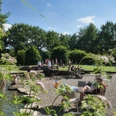 Eine sonnige Parklandschaft mit Personen beim Entspannen und Spielen in einem naturnahen Wasserbereich.