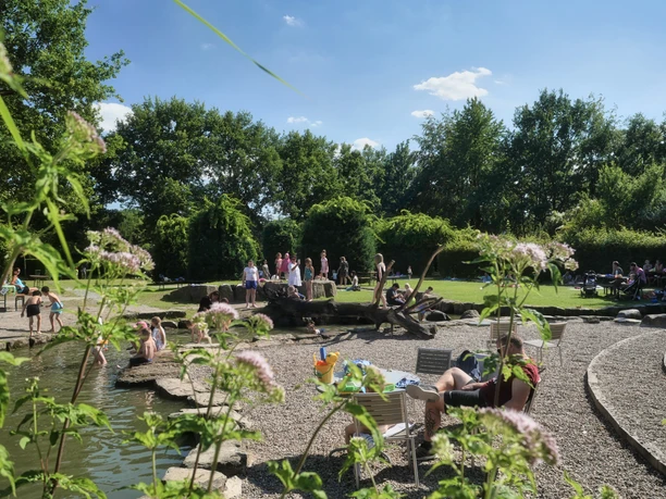 Spielerei im Flora-Westfalica-Park Eine sonnige Parklandschaft mit Personen beim Entspannen und Spielen in einem naturnahen Wasserbereich.
