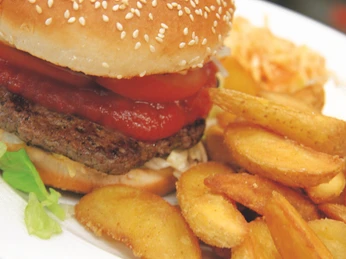 Saftiger Burger mit frischem Salat, dickem Tomato Slice und würzigen Kartoffelecken auf weißem Teller.