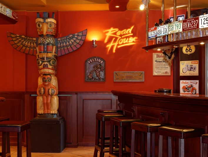 Roadhouse-Innen-03.jpg Eine rustikale Bar im amerikanischen Stil mit Totempfahl und Nummernschildern an roten Wänden.