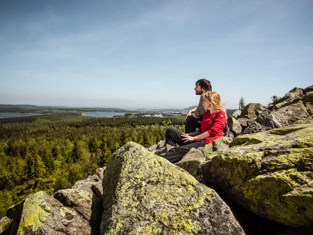 Kammweg Erzgebirge Vogtland Zwei Personen sitzen auf moosbewachsenen Felsen, mit weitem Blick über Wälder und Hügel.