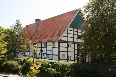 Fachwerkhaus mit rotem Ziegeldach, umgeben von Bäumen und Hecken, bei sonnigem Wetter.