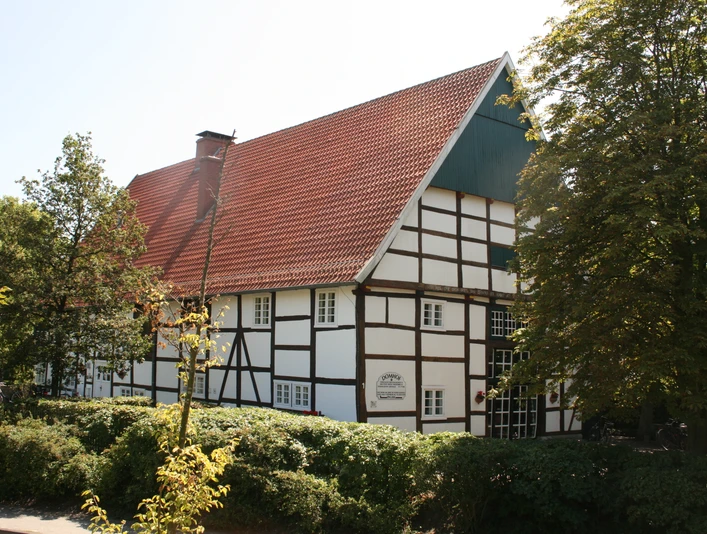 Domhof1.JPG Fachwerkhaus mit rotem Ziegeldach, umgeben von Bäumen und Hecken, bei sonnigem Wetter.