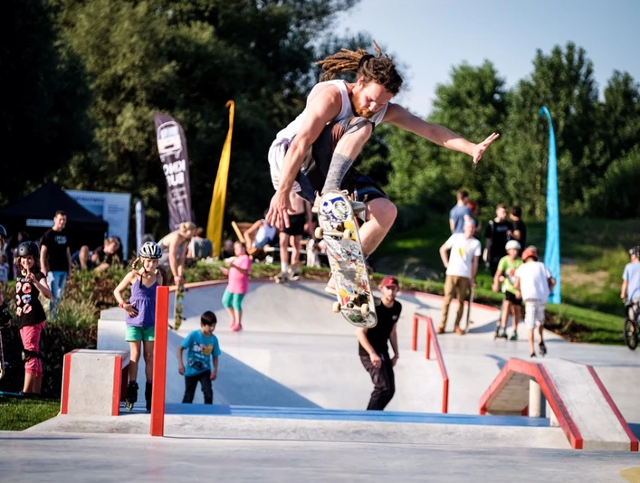 Ein Skateboarder führt in einem lebhaften Skatepark bei sonnigem Wetter einen Sprungtrick aus.