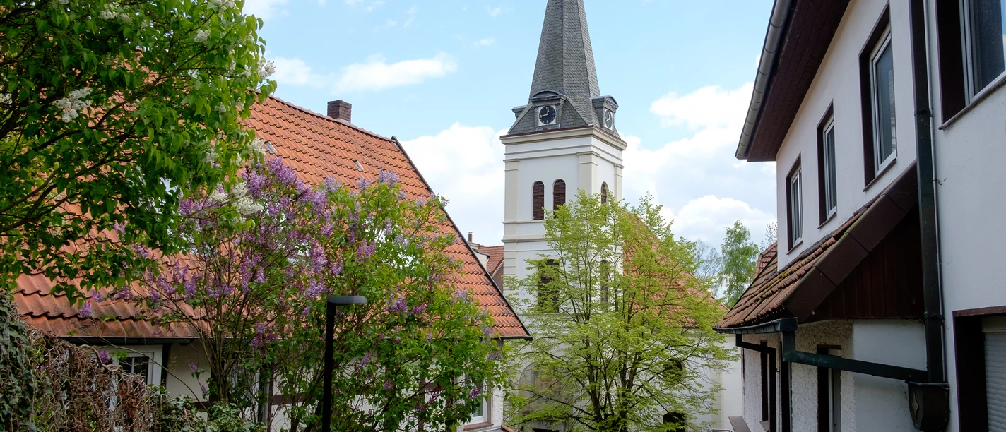 St. Johannis Kirchturm von St. Johannis ragt zwischen rot gedeckten Dächern empor, umgeben von blühenden Bäumen.