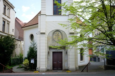 St. Johannis Kleine Backsteinkirche St. Johannis mit Turm, umgeben von Bäumen, auf gepflastertem Platz.