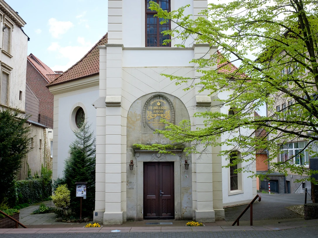 St. Johannis Kleine Backsteinkirche St. Johannis mit Turm, umgeben von Bäumen, auf gepflastertem Platz.