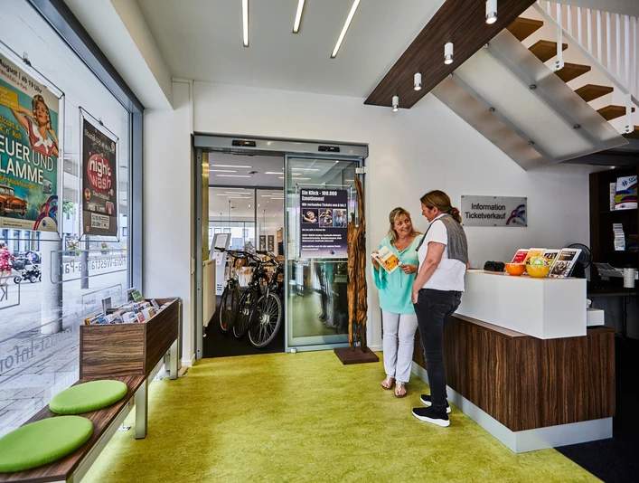 Tourist-Info Rheda-Wiedenbrück Zwei Frauen im modernen Tourist-Info-Büro mit Broschüren und mobilen Fahrrad-Verleihmöglichkeiten.