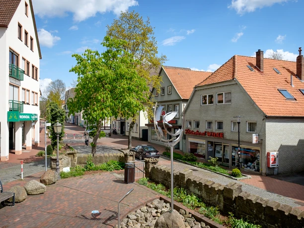 Eine kleine Stadtplatzszene mit modernem Kunstwerk, umgeben von Geschäften und Wohnhäusern.