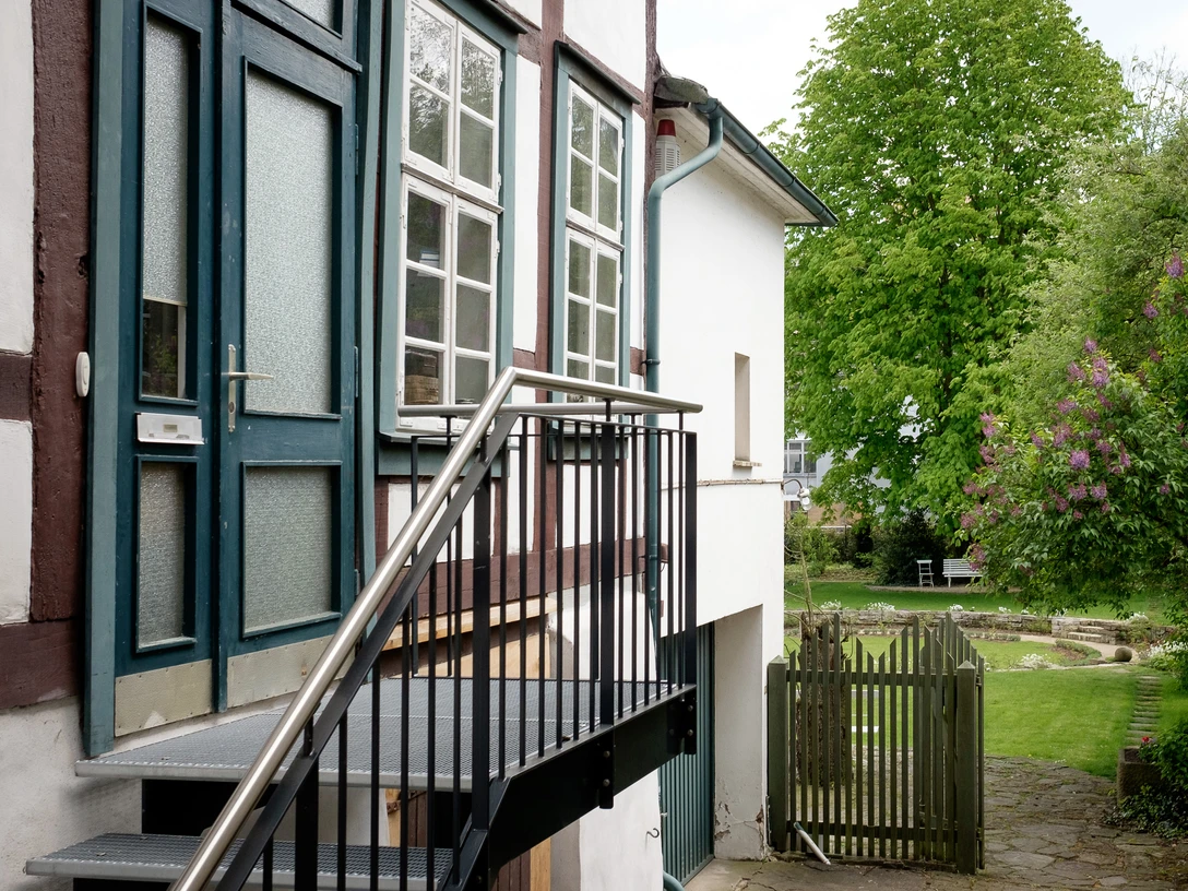 Historische Eingangstreppe führt zu einem zweistöckigen Fachwerkhaus; Garten im Hintergrund mit Flieder.