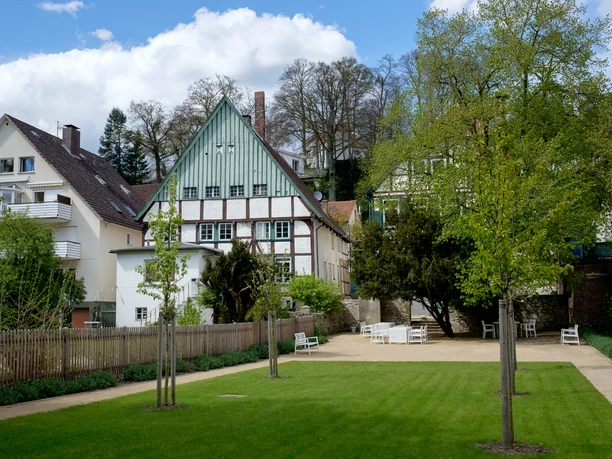 Haus Malz Rückansicht Historisches Gebäude mit grünem Fachwerk, umgeben von Bäumen und gepflegtem Garten mit Parkbänken.