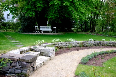 Ein gepflegter Garten mit geschwungenem Kiesweg, Sitzbank und Steinmauer unter dichten Bäumen.