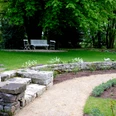 Ein gepflegter Garten mit geschwungenem Kiesweg, Sitzbank und Steinmauer unter dichten Bäumen.