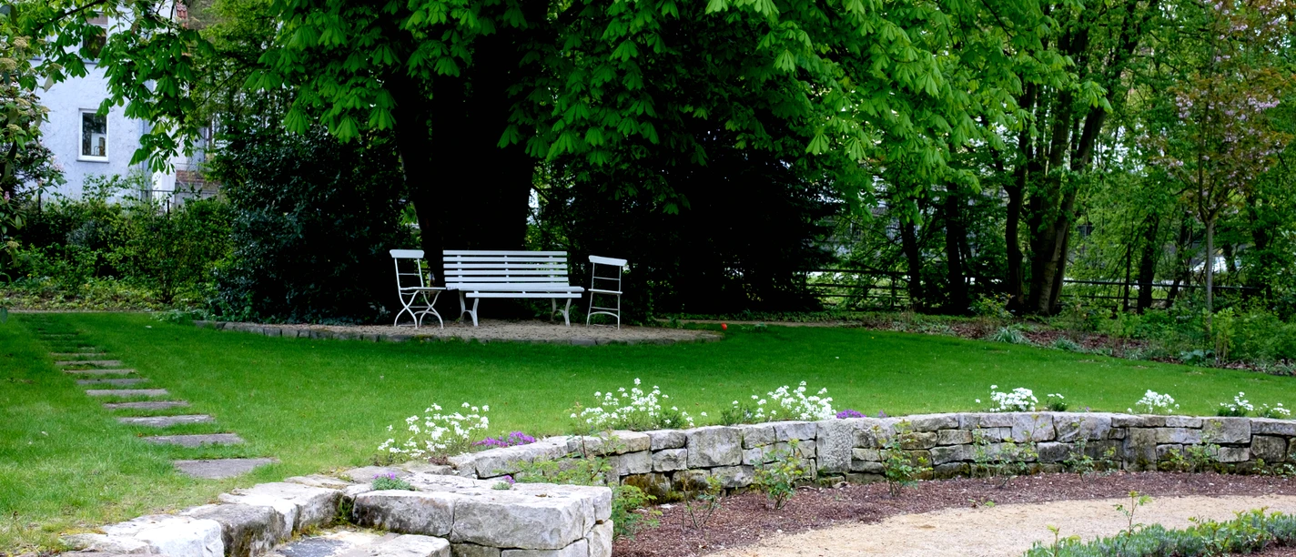 Ein gepflegter Garten mit geschwungenem Kiesweg, Sitzbank und Steinmauer unter dichten Bäumen.