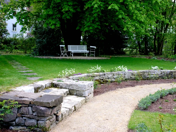 Garten Malz Ein gepflegter Garten mit geschwungenem Kiesweg, Sitzbank und Steinmauer unter dichten Bäumen.