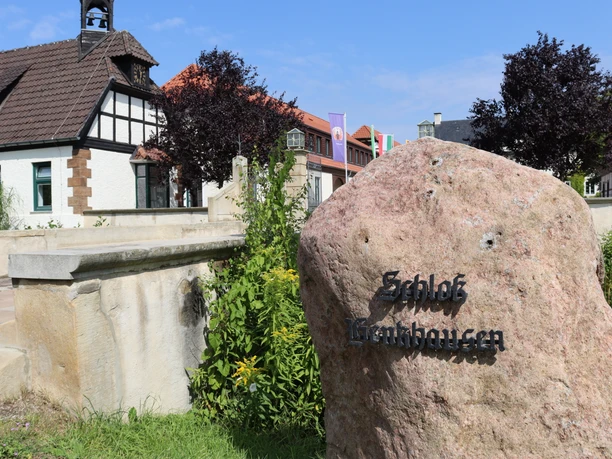 Großer Stein mit der Aufschrift "Schloss Benkhausen" vor traditionellen Fachwerkgebäuden im Hintergrund.
