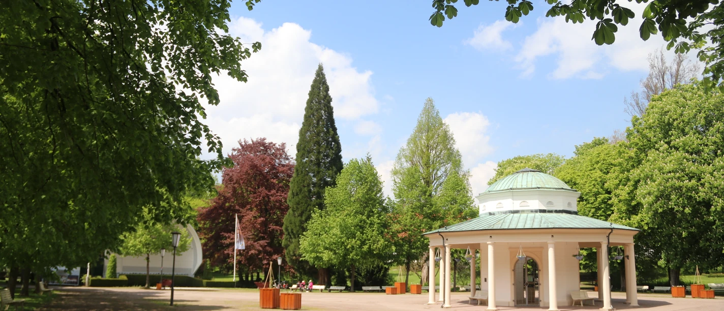 Brunnentempel im Park mit grünem Dach, umgeben von Bäumen und einem klaren, blauen Himmel.