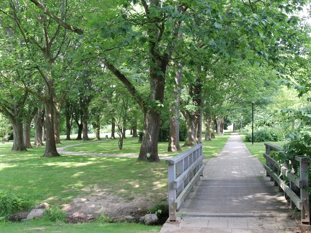 Grüner Park mit Bäumen, Wiesen und einem Fußweg, der über eine kleine Holzbrücke führt.