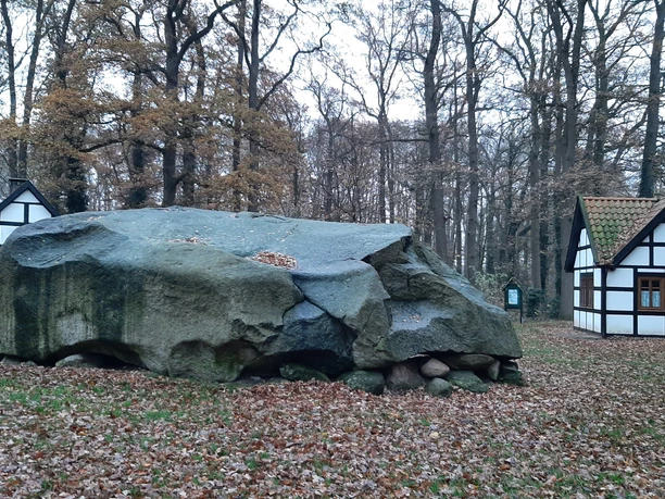 Große Stein Tonnenheide Großer Findling im Wald, flankiert von zwei traditionellen Fachwerkhäusern; Herbstlaub bedeckt den Boden.