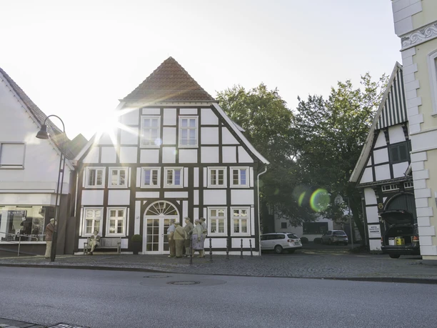 14-08 14-08 Rheda-Wiedenbrück-0300.jpg Fachwerkhaus in Dorfstraße mit Sonnenschein im Hintergrund, Menschen davor, Auto und Bäume daneben.