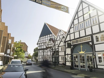 AnkerVilla Fachwerkhäuser und vorbeifahrende Autos in einer sonnigen Straßenansicht, blauer Banner am Himmel.