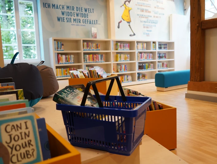 Ein heller Kinderbereich einer Bibliothek mit Bücherregalen, einem blauen Sofa und einem Korb voller Bücher.