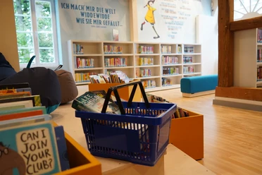 Kinderbibliothek Ein heller Kinderbereich einer Bibliothek mit Bücherregalen, einem blauen Sofa und einem Korb voller Bücher.