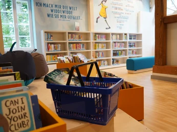 Kinderbibliothek Ein heller Kinderbereich einer Bibliothek mit Bücherregalen, einem blauen Sofa und einem Korb voller Bücher.