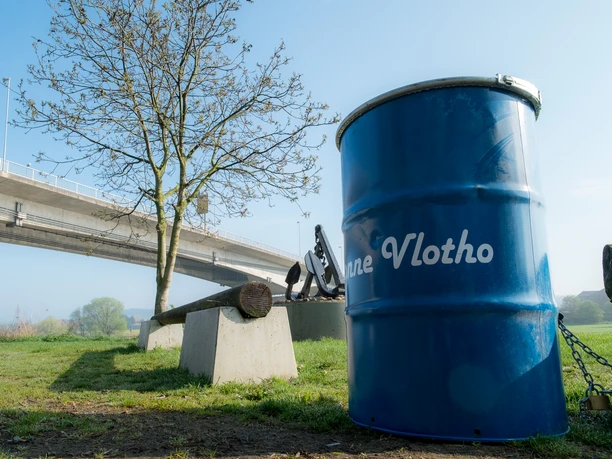 Hafen Vlotho Vorne rechts eine blaue Tonne, dahinter ein Anker auf einem Podest, ganz hinten die WeserbrĂĽcke