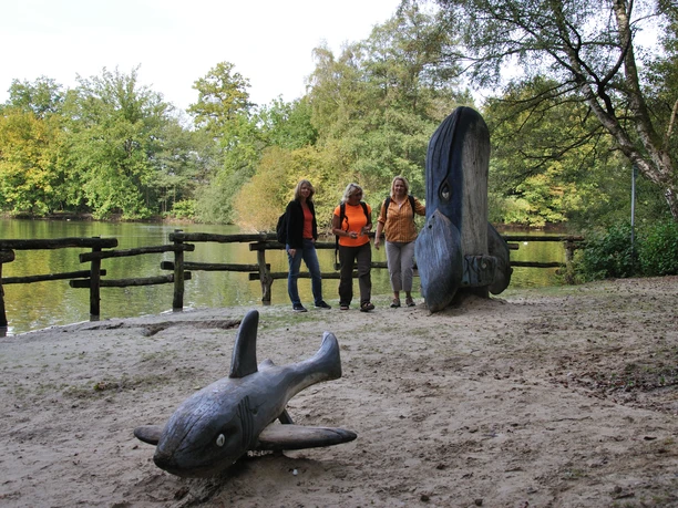 Drei Frauen stehen vor einem Teich nah eines großen Holzkunstwerks in Fischform am Ufer.