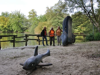 Naherholungsgebiet Fischteiche Drei Frauen stehen vor einem Teich nah eines großen Holzkunstwerks in Fischform am Ufer.