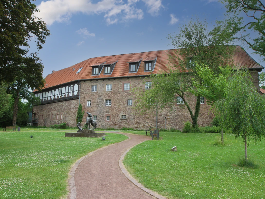 Historisches Gebäude mit rotem Dach, umgeben von grüner Rasenfläche und Gehweg, Skulptur im Vordergrund.