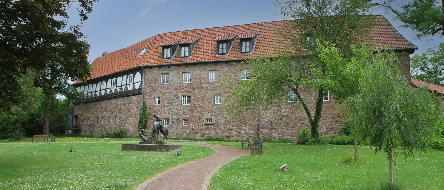 Historisches Gebäude mit rotem Dach, umgeben von grüner Rasenfläche und Gehweg, Skulptur im Vordergrund.