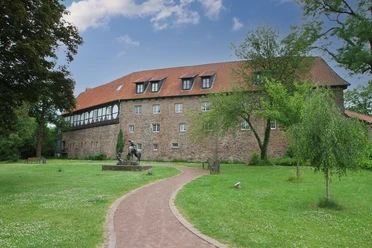 IMG_7929_B.jpg Historisches Gebäude mit rotem Dach, umgeben von grüner Rasenfläche und Gehweg, Skulptur im Vordergrund.